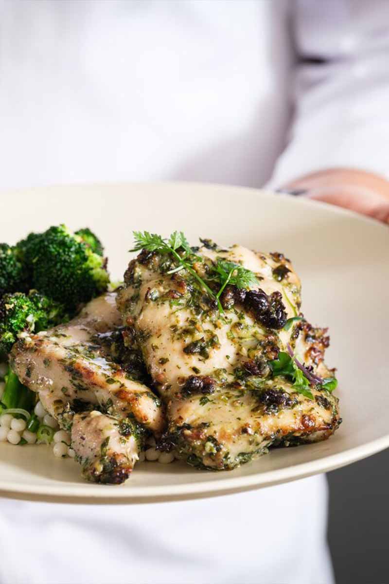 AA person dressed in white is holding a plate with pieces of herb-seasoned grilled chicken and a side of cooked broccoli.