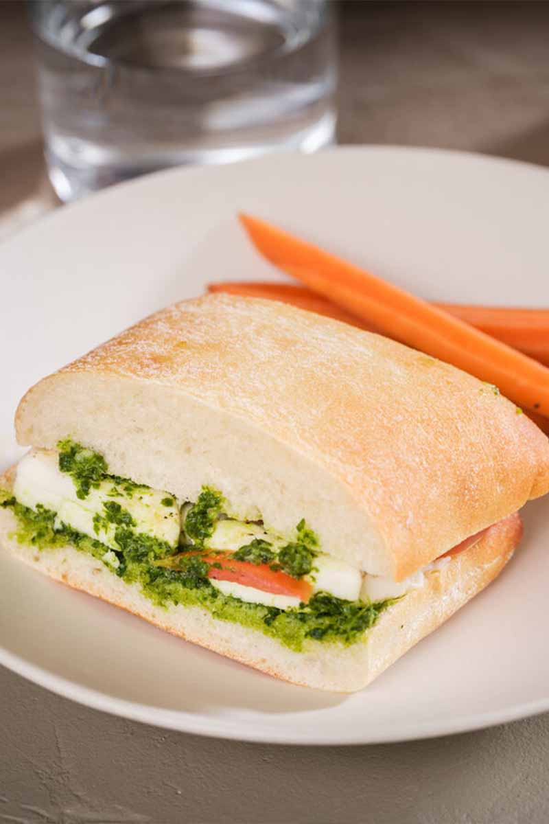 A sandwich with green pesto sauce and cheese on a white plate, accompanied by two baby carrots, set on a table with a glass of water and utensils nearby.