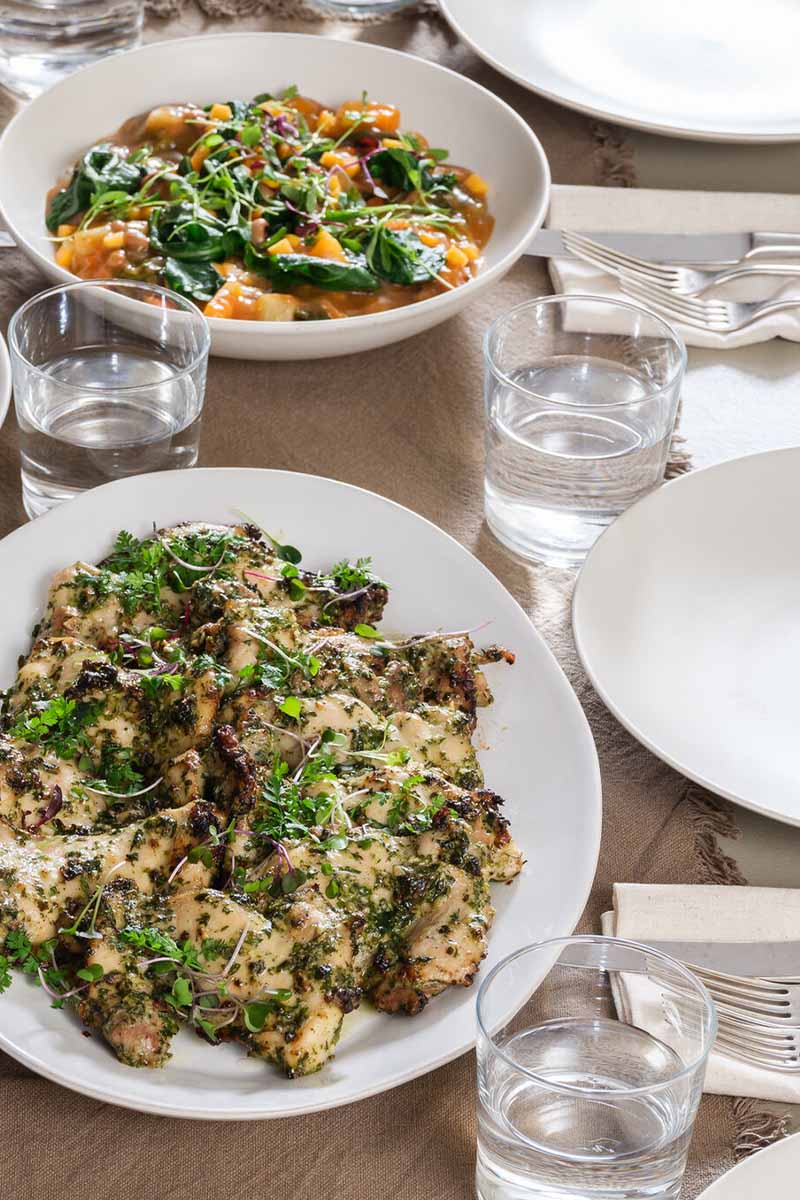 A dining table is set with empty white plates, glasses of water, and utensils, accompanied by bowls of prepared dishes including a greens salad, a pasta or grain dish, and another mixed vegetable dish, in a cozy indoor setting.