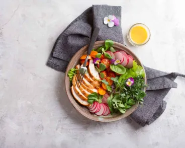 A bowl of fresh salad with grilled chicken slices, radishes, cherry tomatoes, greens, and edible flowers, accompanied by a fork and a small glass of orange juice on a gray napkin, set on a light-colored surface.