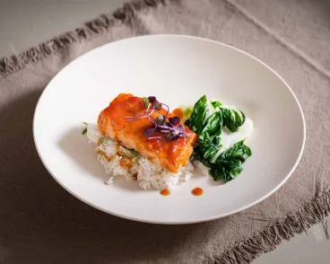 A plated dish featuring a piece of glazed salmon topped with microgreens, served on a bed of white rice alongside a portion of cooked leafy greens, all arranged on a white plate set on a beige textured cloth.