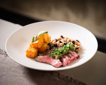 A white plate with sliced medium steak topped with green herbs, accompanied by cubed roasted butternut squash and a serving of rice mixed with black beans, all set on a gray cloth surface.