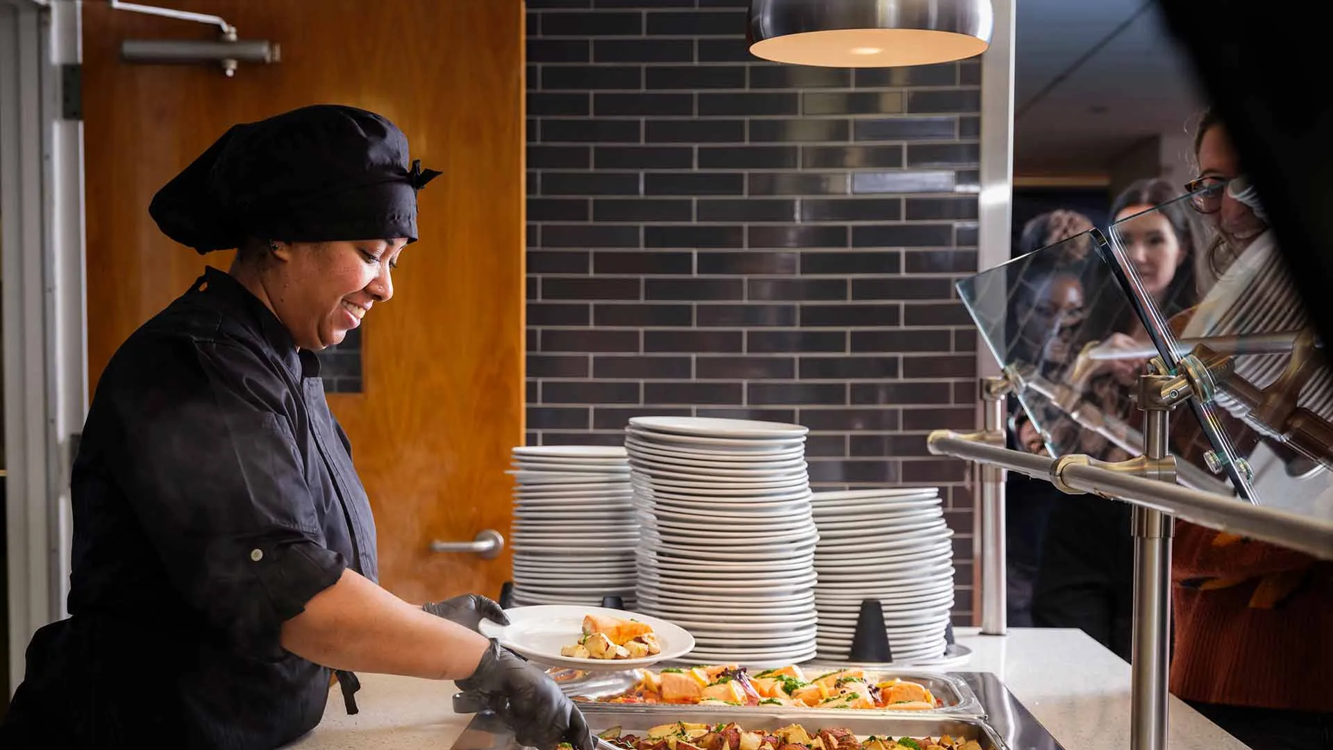 A chef wearing a black uniform and hat is smiling while serving food from trays onto plates for people waiting in line at a buffet-style counter.