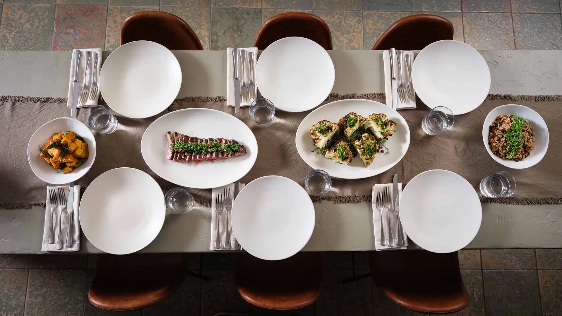 A dining table set for six people with plates, utensils, and glasses, alongside dishes of food including a fish fillet, stuffed artichokes, meat with garnish, and roasted vegetables arranged in a casual dining setting.