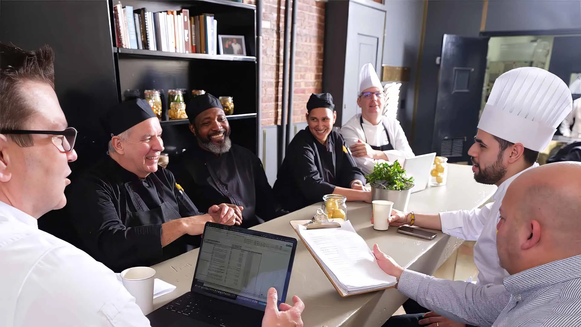 A group of chefs and two men in casual business attire are seated around a kitchen counter, engaged in a lively discussion with one man holding a laptop and another showing notes on a clipboard in a professional kitchen setting.