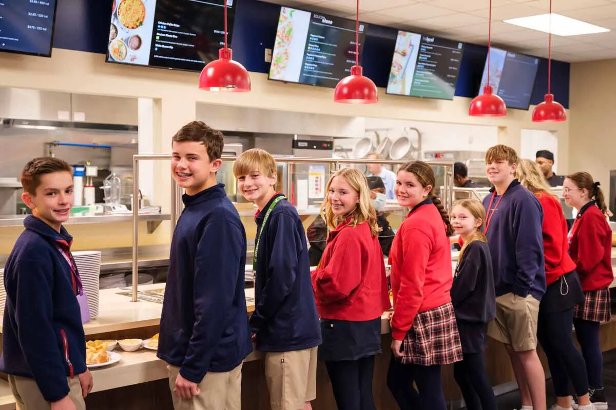 A group of smiling school-aged children stand in line in a brightly lit cafeteria, waiting to receive food from the serving counter.