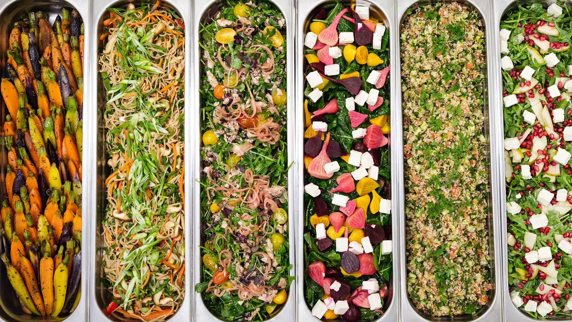 An overhead view of a salad bar with eight metal trays filled with a variety of fresh, colorful salads and vegetables, set in a restaurant or buffet setting.