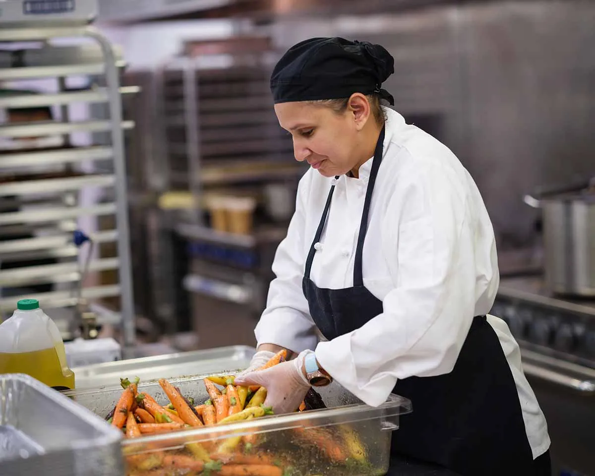 A chef in a professional kitchen, wearing a white coat, black apron, and headscarf, washing and preparing a large container of fresh carrots.