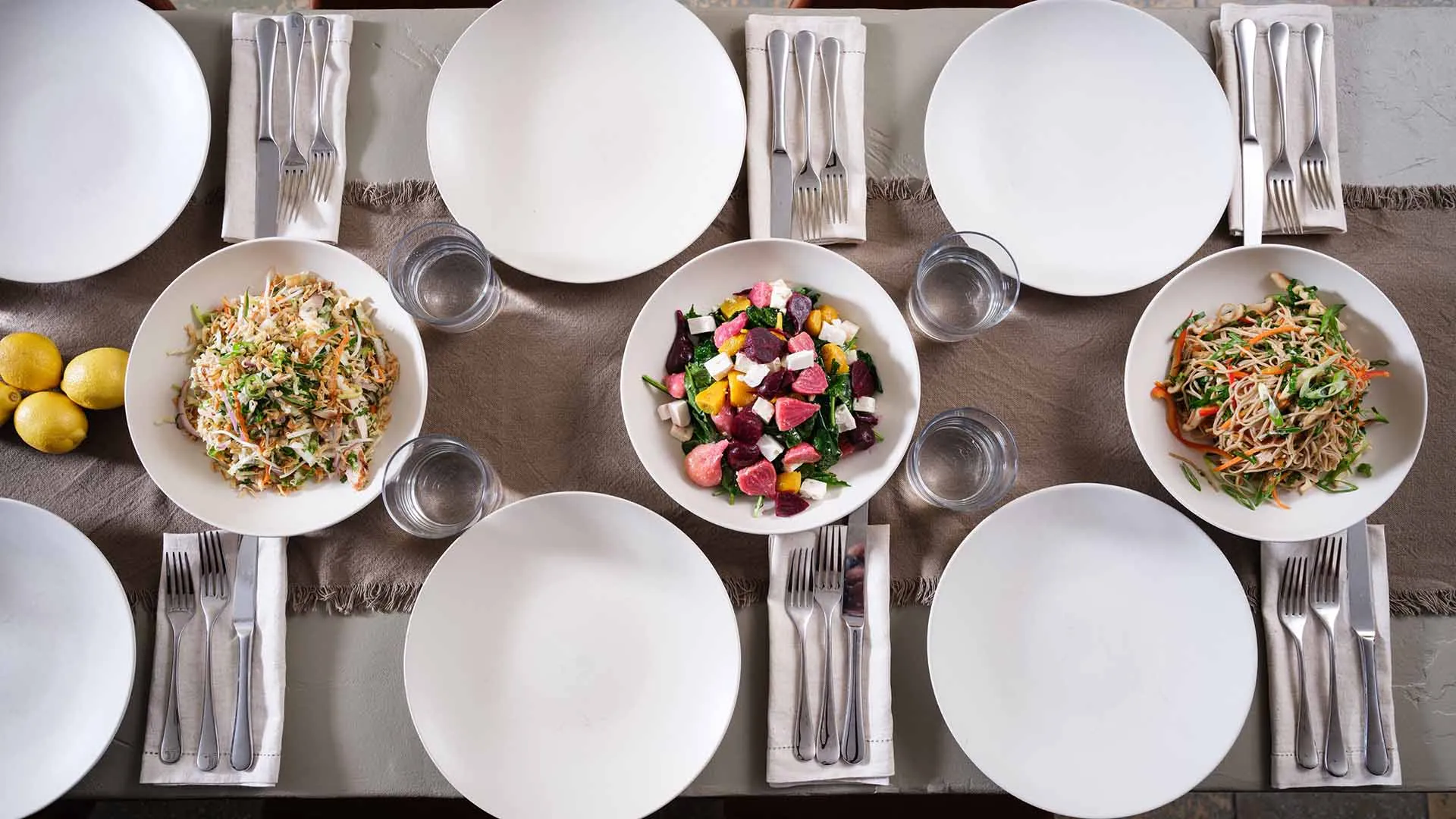 A neatly set dining table with six place settings, each having a plate, cutlery, and a glass, along with three bowls of colorful salads placed in the center of the table.