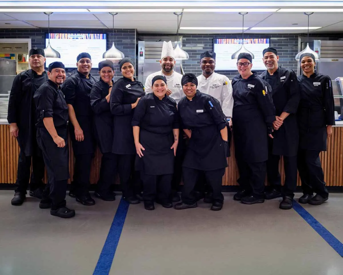 A diverse group of eleven chefs and kitchen staff, dressed in black uniforms and white chef hats, pose together and smile confidently in a modern commercial kitchen.
