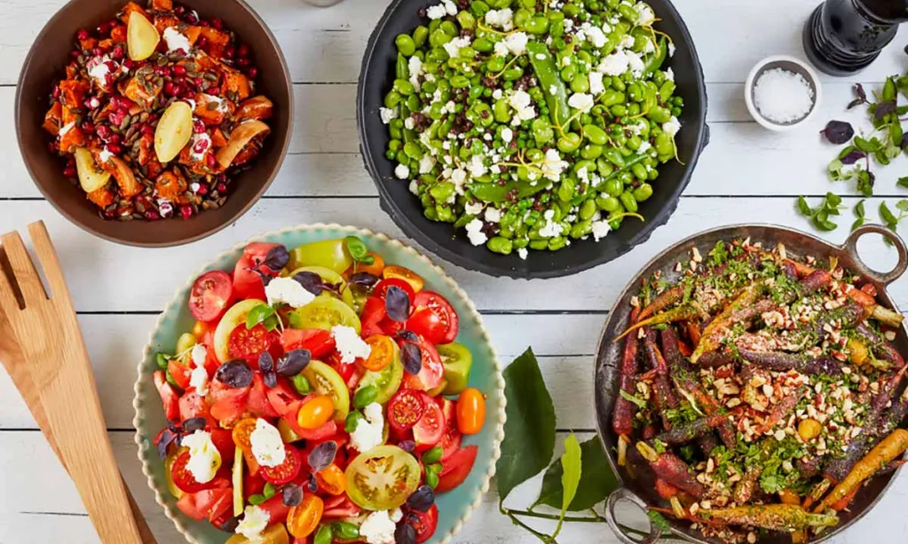 A colorful spread of four fresh vegetable dishes arranged on a white wooden table, accompanied by seasonings and wooden serving utensils.