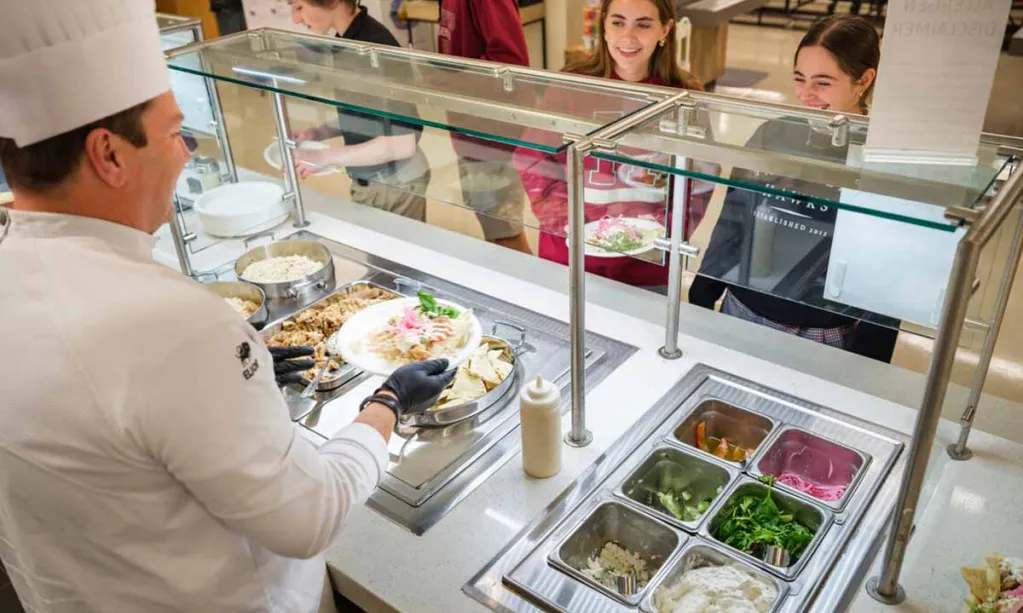 A chef in a white uniform is serving food to smiling students standing behind a cafeteria counter in a school dining area.
