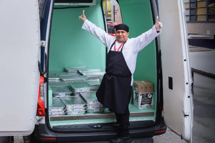 A chef happily standing with thumbs up and smiling near the back of a delivery van full of pans of food