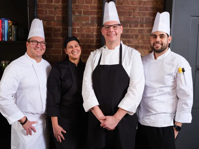 Four smiling chefs dressed in professional kitchen attire standing together in a restaurant or kitchen setting with a brick wall background.