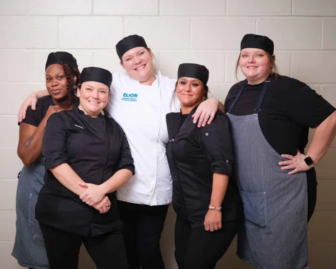 Five smiling chefs wearing black hats and uniforms, standing closely together and posing in front of a plain white brick wall.