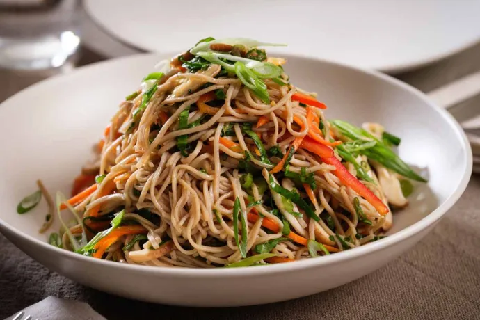 a plate of noodles mixed with sliced vegetables, such as carrots and green onions, served in a white bowl on a table.