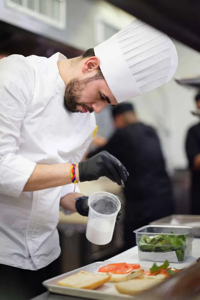 A chef in a white uniform and hat is carefully seasoning or sprinkling an ingredient over open sandwiches in a professional kitchen setting.