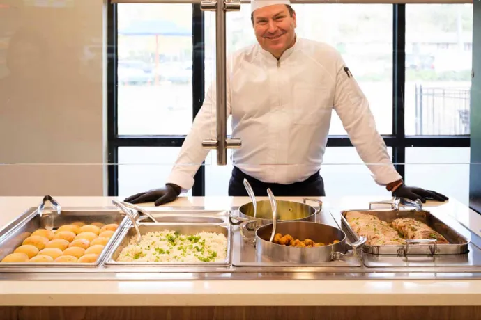 A chef in a white uniform and hat is standing behind a food counter with trays of prepared dishes in a well-lit, modern cafeteria setting.