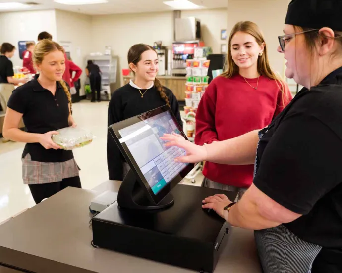 Three young women at a cafeteria counter, with one working behind the register and the others interacting as customers.