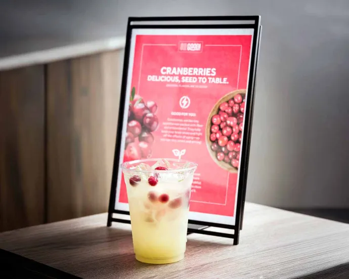 A clear plastic cup filled with a beverage with cranberries in the foreground, set on a wooden surface with a informational sign about cranberries in the background.