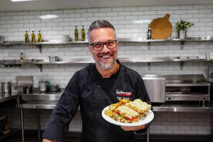 A smiling chef in a black uniform holding a plate of food with sandwiches and potato chips in a modern kitchen setting.